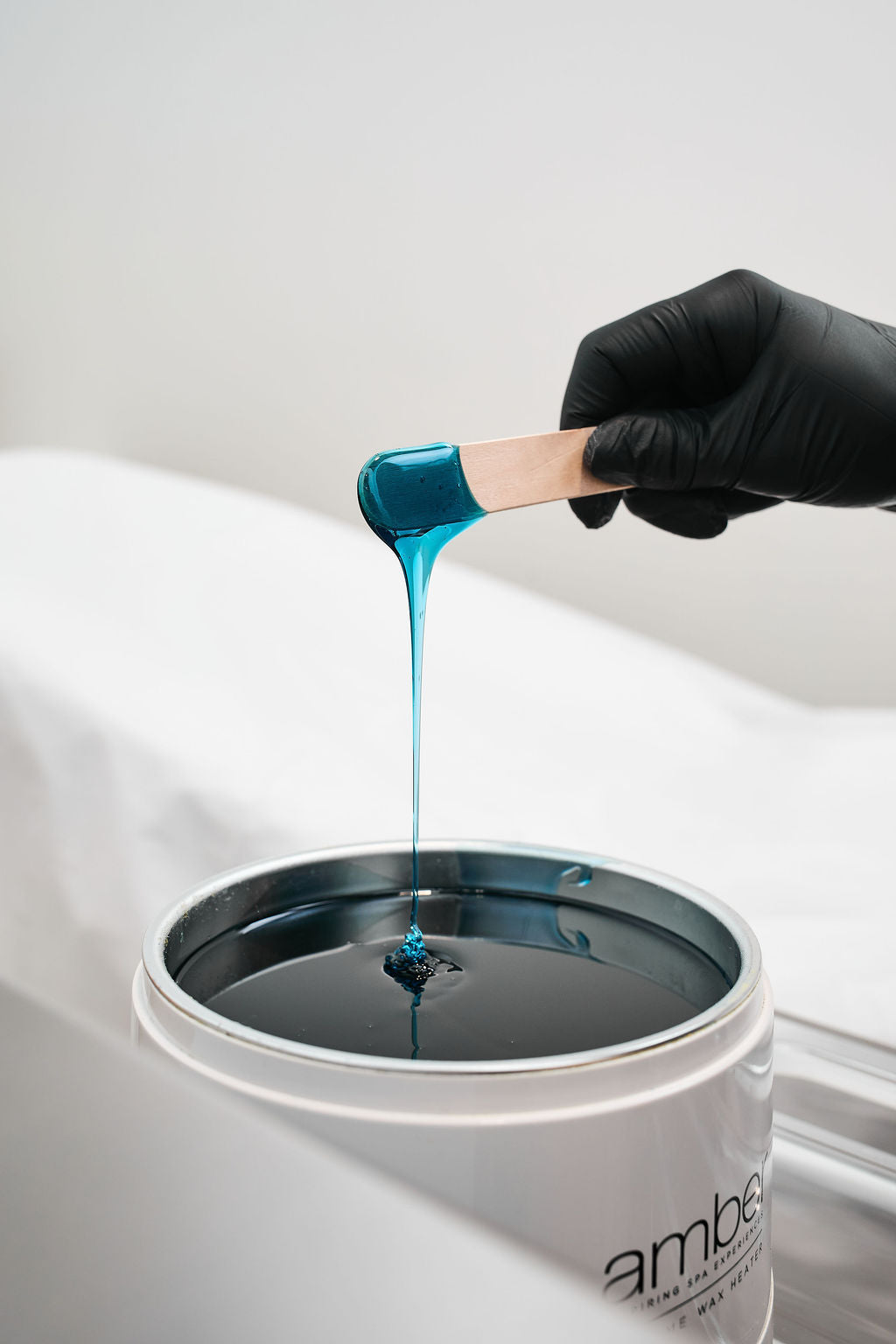 Hand wearing a black glove dipping a wooden stick into a container of blue liquid, with 'ambesoin' branding visible.